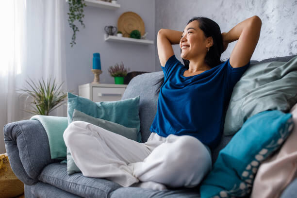 Full length shot of beautiful, happy young woman sitting on the cozy sofa with hands behind head, looking away, smiling and daydreaming.